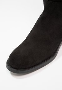 Svart mockakänga med en slät yta, rund tå och en kontrasterande svart gummisula med fina sömmar längs kanten.