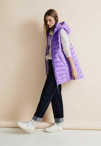 Lila pufferväst med huva, med horisontell sömnad, framfickor med dragkedja, kombinerad med mörka jeans och vita sneakers.