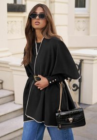 Haine tip poncho de culoare neagră cu margini albe, strânsă în talie. Combinate cu blugi de culoare albastru deschis și o geantă mică de mână cu imprimeu de crocodil negru.