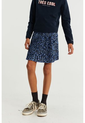 Personne portant un sweat-shirt bleu marine « TRÈS COOL », une jupe plissée à imprimé léopard bleu, des chaussettes noires et des baskets à imprimé léopard, debout sur un sol blanc.