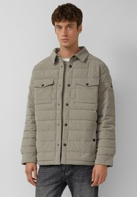 Lichtgrijze gewatteerde jas met een corduroy textuur, knoopsluiting aan de voorkant en twee borstzakken met zwarte knopen.