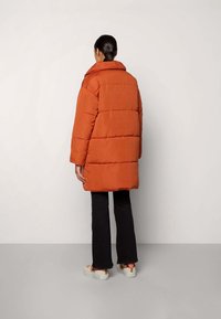 Orange vadderad jacka med hög krage, utrustad med horisontell sömnad och två sidofickor, kombinerad med svarta utsvängda byxor och avslappnade skor.