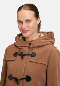 Bruine wollen jas met een capuchon, voorzien van zwarte togglesluiting en een gestructureerde afwerking. De binnenvoering toont een geruit patroon in gedempte kleuren.