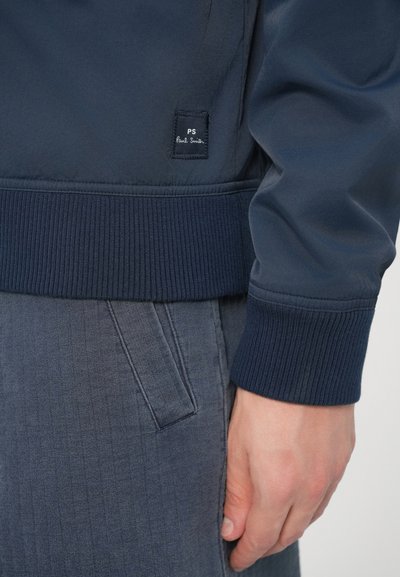 Námornícka modrá bomber bunda s rebrovanými manžetami a lemom. Na boku je malá čierna nášivka s výšivkou "PS" a "Paul Smith".