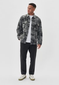 Jachetă de flanel texturată, gri și neagră, cu închidere cu nasturi, două buzunare pe piept, purtată peste un tricou alb, asortată cu pantaloni negri și adidași.