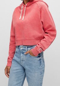 Persona vestida con un hoodie cortado de color coral desgastado, con cordones blancos y vaqueros de cintura alta de color azul claro, con una mano en el bolsillo.