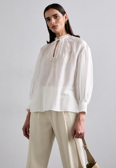 Blouse blanche texturée à manches longues avec un col froncé et un détail à lien, associée à un pantalon clair et ample et un sac beige.