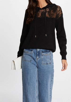 Femme portant un sweat à capuche noir avec un empiècement en dentelle et un jean bleu, tenant une pochette blanche matelassée sur fond blanc.