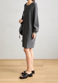 Robe en maille grise légère avec manches larges et poches latérales. Associée à des sandales à talons blocs ouvertes noires. Décor neutre et sol.