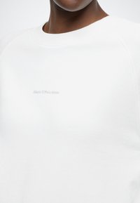 Fehér pulóver puha anyagból, környakkivágással, raglán ujjakkal és a mellrészen dombornyomott "Marc O'Polo DENIM" logóval.