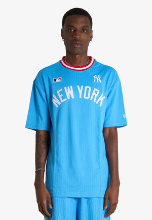 Joven con el pelo corto que lleva una camiseta de béisbol azul de los New York Yankees y pantalones cortos a juego sobre un fondo blanco.