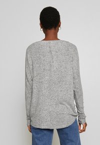 Osoba z krótkimi kręconymi włosami, nosząca złote kolczyki w kształcie kół, sweter z długim rękawem w jasnoszarym melanżu oraz niebieskie dżinsy, stojąca tyłem do aparatu.