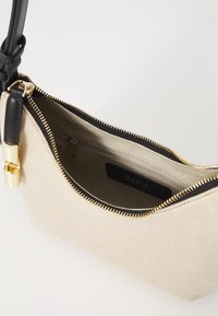 Μπεζ υφασμάτινη τσάντα χεριού με μαύρη δερμάτινη λουρί και χρυσές λεπτομέρειες. Ο εσωτερικός χώρος περιλαμβάνει μια τσέπη με φερμουάρ και ετικέτα επωνυμίας. Κλείσιμο με φερμουάρ.