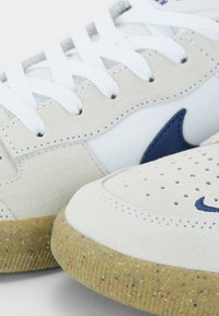Baskets blanches et beiges avec lacets blancs, logo bleu, bout perforé et semelle en caoutchouc translucide mouchetée en gros plan.
