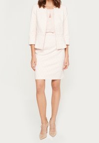 Ensemble tailleur rose pâle comprenant un blazer texturé à manches trois-quarts, une jupe assortie et des talons nus à lanières. Détail en forme de goutte au niveau du col.