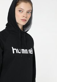 Svart hoodie med en framövlig logotyp i vitt, med en justerbar huva och ett texturerat innerfoder. Enkel, sportig design.
