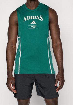 Svalnatá osoba s zeleným bezrukávovým tréninkovým topem Adidas s bílým logem a černými sportovními šortkami stojící před jednobarevným pozadím.