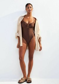 Bañador marrón de una sola pieza con escote fruncido, llevado debajo de una camisa abierta de color beige claro. Se presentan sandalias planas con un detalle de hebilla.