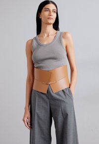 Ceinture en cuir brun avec un design angulaire, associée à un débardeur en jersey gris à côtes et à un pantalon gris ample, créant un look superposé.