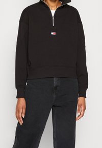 Felpa nera con zip a quarto, dettagli geometrici nelle cuciture e un piccolo logo rosso, bianco e blu sul petto.
