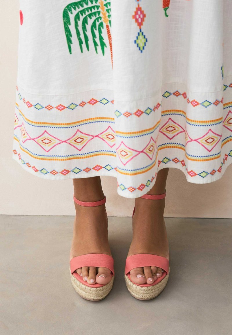 Sandales roses à lanières avec une semelle espadrille, associées à une robe blanche brodée ornée de motifs géométriques colorés et de motifs de palmiers.