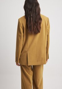 Blazer jaune moutarde avec épaulettes, fente unique à l'arrière et poignets boutonnés, associé à un pantalon large assorti.
