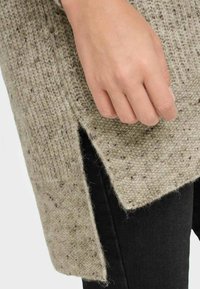 Hellgrauer Strickpullover mit einem strukturierten Muster und seitlichem Schlitz, kombiniert mit schwarzen Hosen. Die Hand ruht auf dem Rand des Pullovers.