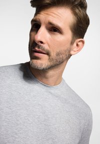 Szara bawełniana koszulka z okrągłym dekoltem i subtelnym tłoczonym logo na piersi. Gładka textura, dopasowany krój, krótkie rękawy.