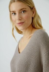 Pull à col en V en maille côtelée gris clair, avec une coupe décontractée et une texture douce. Le modèle a les cheveux blonds ondulés et porte peu d'accessoires.