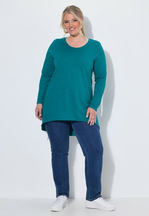 Lachende vrouw die een teal gekleurde tuniek met lange mouwen, blauwe spijkerbroek en witte sneakers draagt, staand tegen een effen lichte achtergrond.