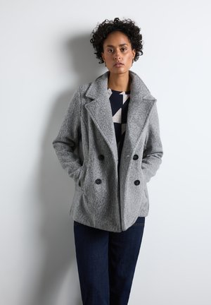 Cappotto grigio doppiopetto in lana strutturata, con grandi bottoni neri, risvolti larghi e tasche laterali, indossato sopra una maglietta a fantasia.