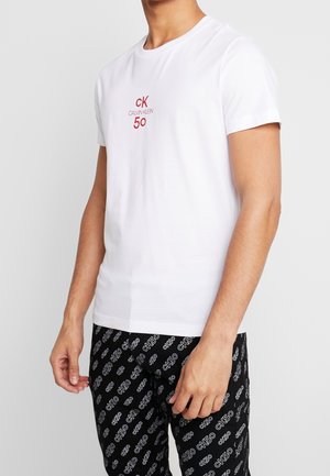 T-shirt en coton blanc avec un logo rouge "CK CALVIN KLEIN 50" sur le devant ; porté avec un pantalon de pyjama noir affichant un motif textuel répétitif.
