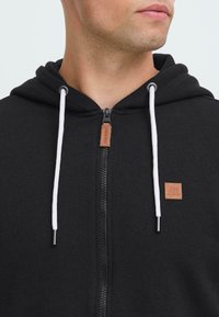 Uomo che indossa una felpa nera con zip, con stringhe bianche e toppe in pelle marrone sulla cerniera e sul petto.