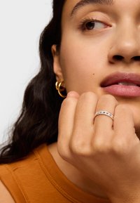 Anello d'argento con piccole pietre chiare, indossato su una mano con tonalità di pelle naturale. Orecchino a cerchio dorato visibile. La modella indossa una maglia color ruggine.