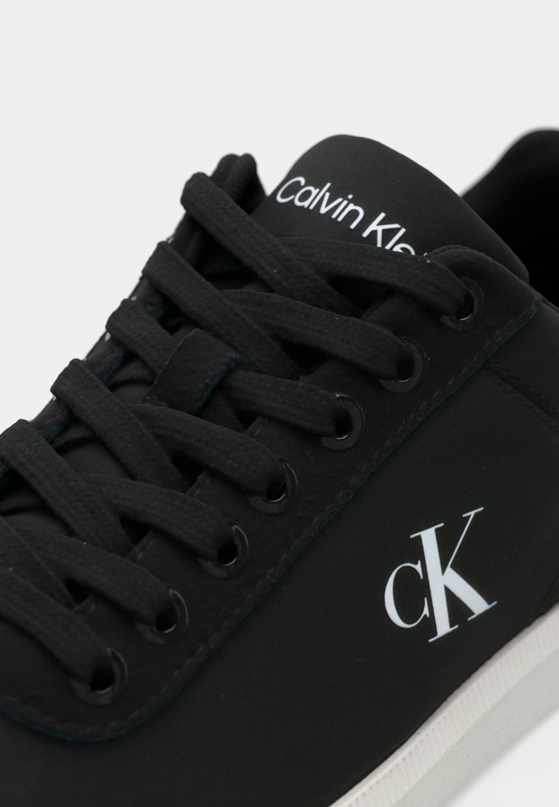 Black sneaker with round toe, smooth synthetic material, flat laces, and a white Calvin Klein logo on the side. Rubber sole and textured details.