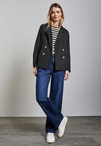 Chaqueta negra de doble botonadura con botones plateados, llevada sobre una camiseta de rayas negras y blancas, combinada con jeans de pierna ancha azules y zapatillas blancas.