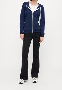 Navy zip-up hoodie with white drawstrings, front pockets, paired with black flared pants and silver-accented sneakers. Smooth fabric texture.