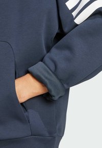 Marineblauwe hoodie met een zachte textuur, voorzien van een voorkantzak en opgerolde manchetten. De display bevat witte accentstrepen op de mouwen.