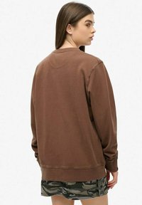 Felpa marrone con polsini e orlo a costine, caratterizzata da un classico scollo tondo. Indossata sopra pantaloncini mimetici, che mettono in risalto una vestibilità comoda.