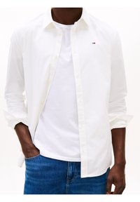 Camisa blanca abotonada sobre una camiseta blanca de cuello redondo, combinada con jeans de mezclilla azul. Tiene un pequeño logo en el pecho de la camisa.