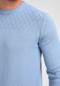 Zbliżenie na osobę w jasnoniebieskim swetrze z dzianiny, posiadającym diamentowy i poziomy prążkowany wzór na klatce piersiowej i rękawach.