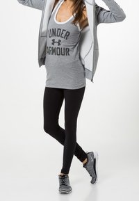 Camiseta sin mangas gris con "UNDER ARMOUR" en negrita y color negro, combinada con leggings negros y zapatillas texturizadas en negro y gris. Chaqueta gris con capucha.