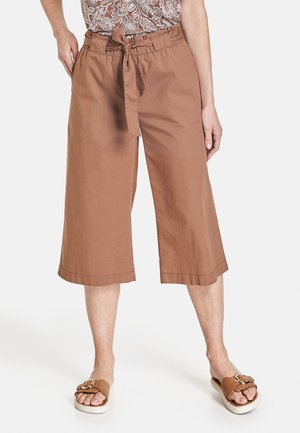 Femme portant un pantalon marron large et court avec une ceinture nouée à la taille et des sandales marron avec des détails de boucle dorée.