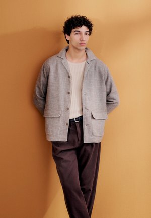 Jauns vīrietis ar cirtainiem matiem, atspiedies pret sinepju krāsas sienu, ģērbies bēša adītā džemperī, gaiši pelēkā pogātas žaketes un tumši brūnās biksēs.