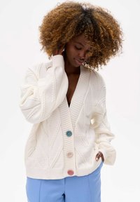 Cream cable-knit cardigan featuring a deep V-neck, three buttons in blue, pink, and green, and loose-fitting, ribbed cuffs and hem.