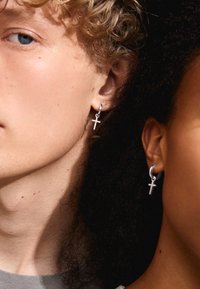 Pendientes de aro de plata con pequeños charms de cruz, llevados por una persona con cabello rizado y otra con cabello texturizado, mostrando un diseño moderno.