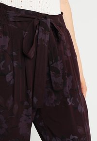 Pantalons bordeaux avec imprimé floral, ceinture en tissu et taille smockée. Présente une texture douce avec des motifs floraux violet fonc é et noir.