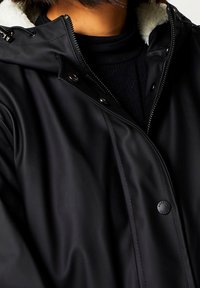 Schwarze wasserdichte Jacke mit einem Reißverschluss vorne, Druckknopfverschluss und einem weichen Fleece-Futter am Kragen; glatte Textur.