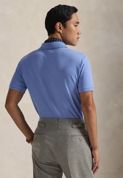 Polo azul claro com acabamento texturizado, com gola azul-marinho com estampa de bolinhas. Calças cinzentas com bolsos traseiros e detalhes em metal dourado.