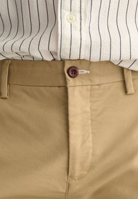 Beige bomulds khaki bukser har en brun knap, flad front og subtil tekstur, parret med en stribet hvid og marineblå skjorte.